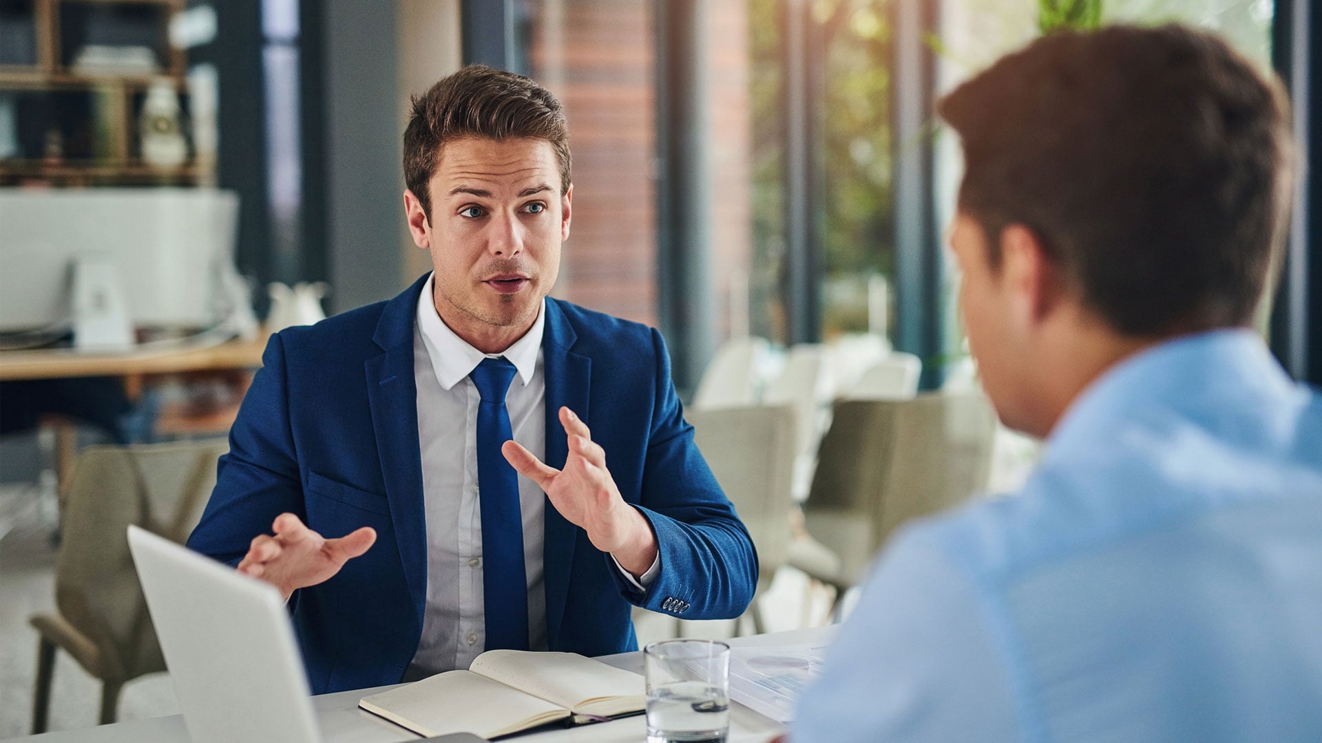 Two men sitting face to face in the office, one with his back to the camera, the other in a navy suit